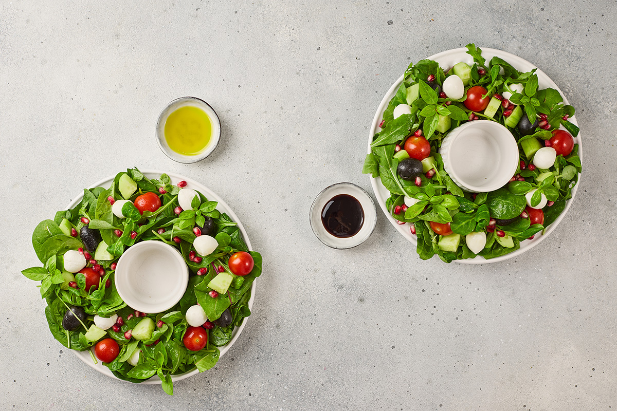 Two Christmas wreath salads side by side with small bowls of balsamic glaze and olive oil ready for serving.