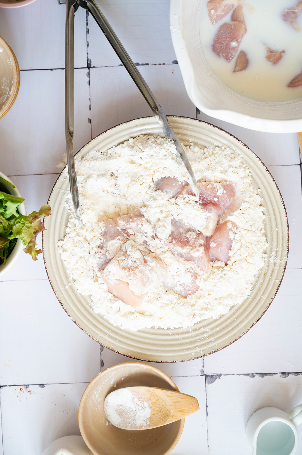 Chicken pieces being coated in a seasoned flour and cornstarch mixture using tongs.