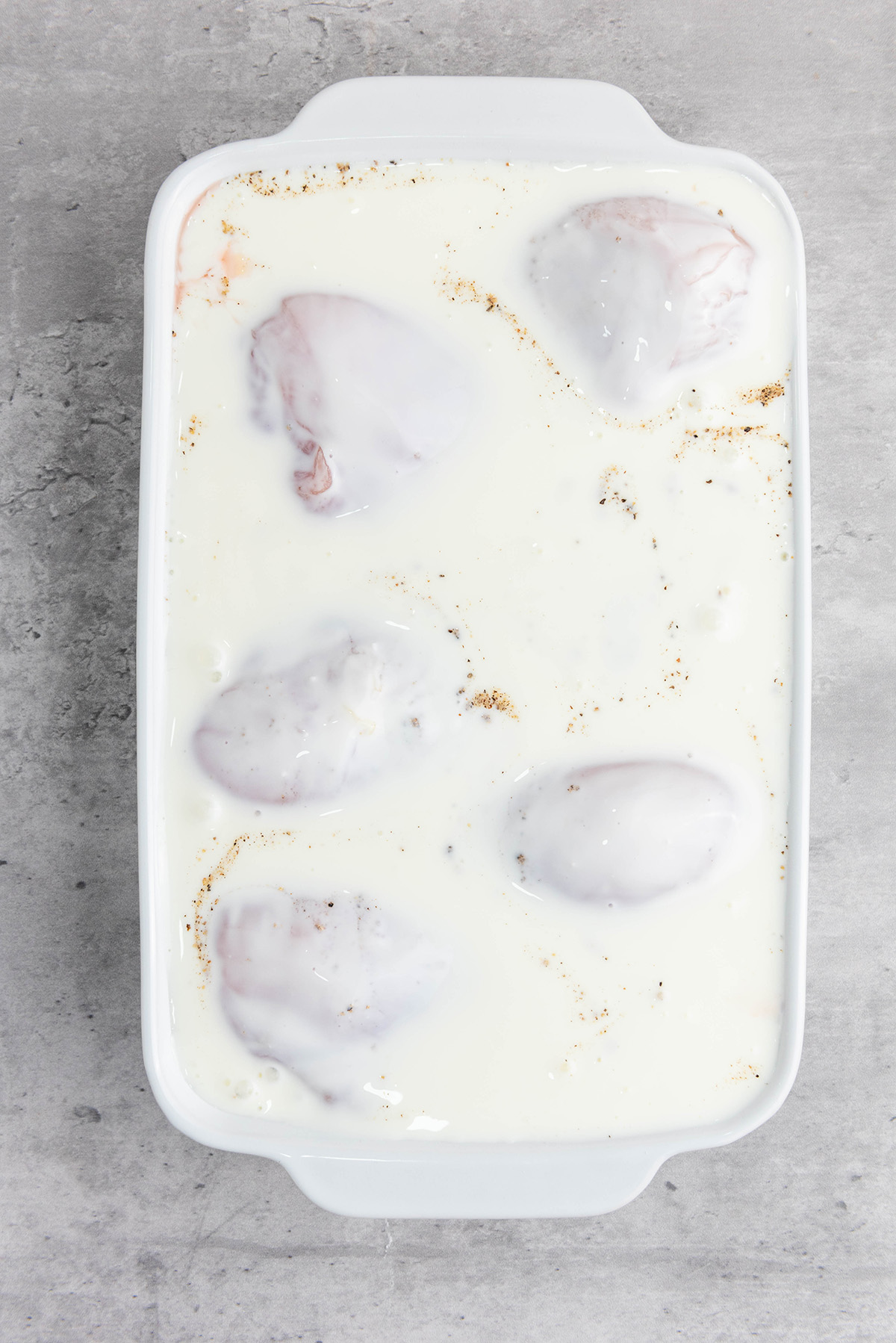 Raw chicken covered in buttermilk marinade in a white baking dish.