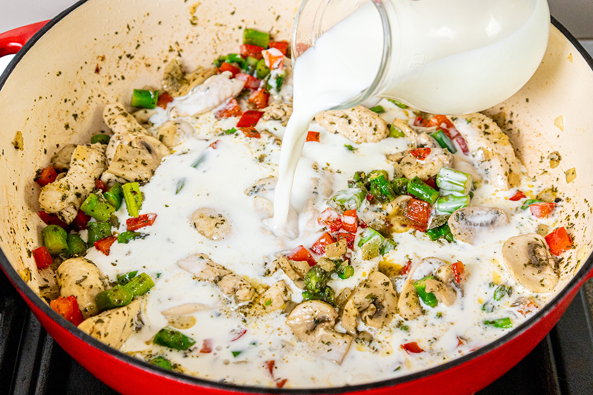 Milk being poured into the skillet with cooked chicken and vegetables to make the creamy alfredo sauce.