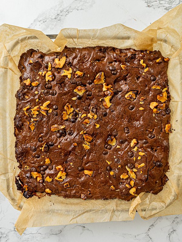 Freshly baked chocolate walnut brownies cooling in a square pan lined with parchment paper.