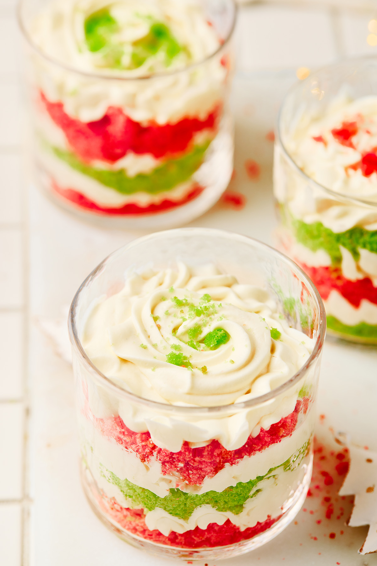 A fun and festive Christmas trifle dessert made with crumbled red and green cakes and an easy, fluffy cream cheese filling that tastes like the holidays in every bite. Close-up of layered red and green Christmas trifles with swirls of cream on top.