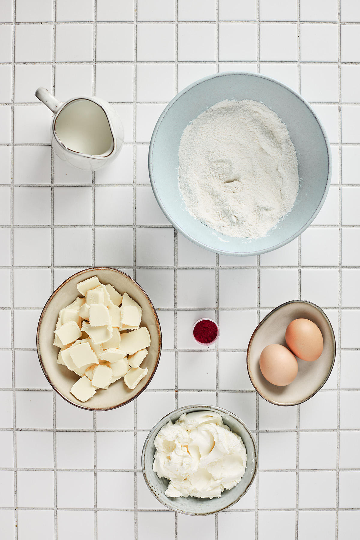 Overhead view of the ingredients for red velvet cake truffles laid out on a white tiled surface, including flour, cubed butter, eggs, milk, red food coloring, and cream cheese frosting.
