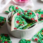 Pieces of Grinch bark with green swirls, red candy hearts, and colorful sprinkles piled in a white ramekin with extra pieces scattered around.