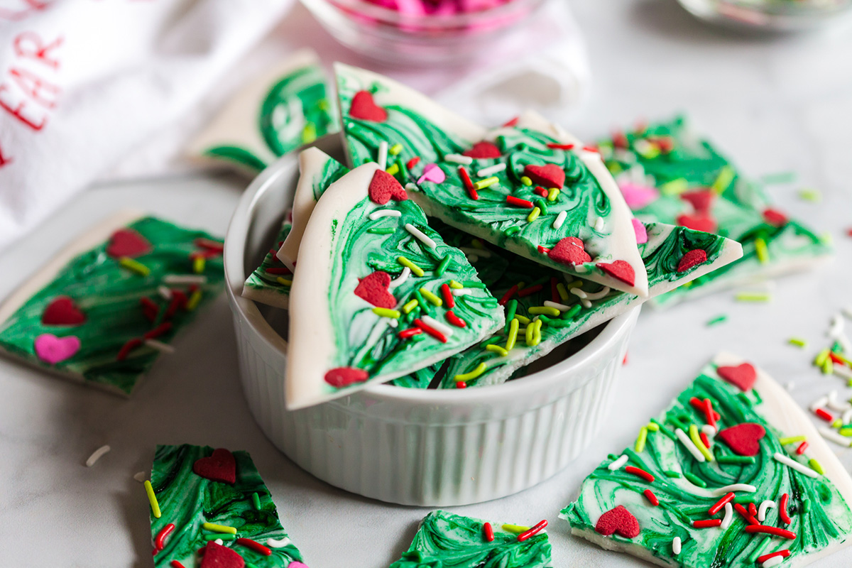 Pieces of Grinch bark with green swirls, red candy hearts, and colorful sprinkles piled in a white ramekin with extra pieces scattered around.