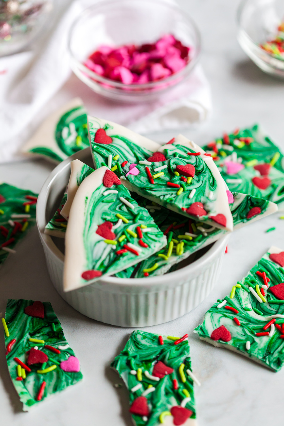 Triangular pieces of Grinch bark with green swirls and festive sprinkles stacked in a white ramekin with bowls of sprinkles in the background.
