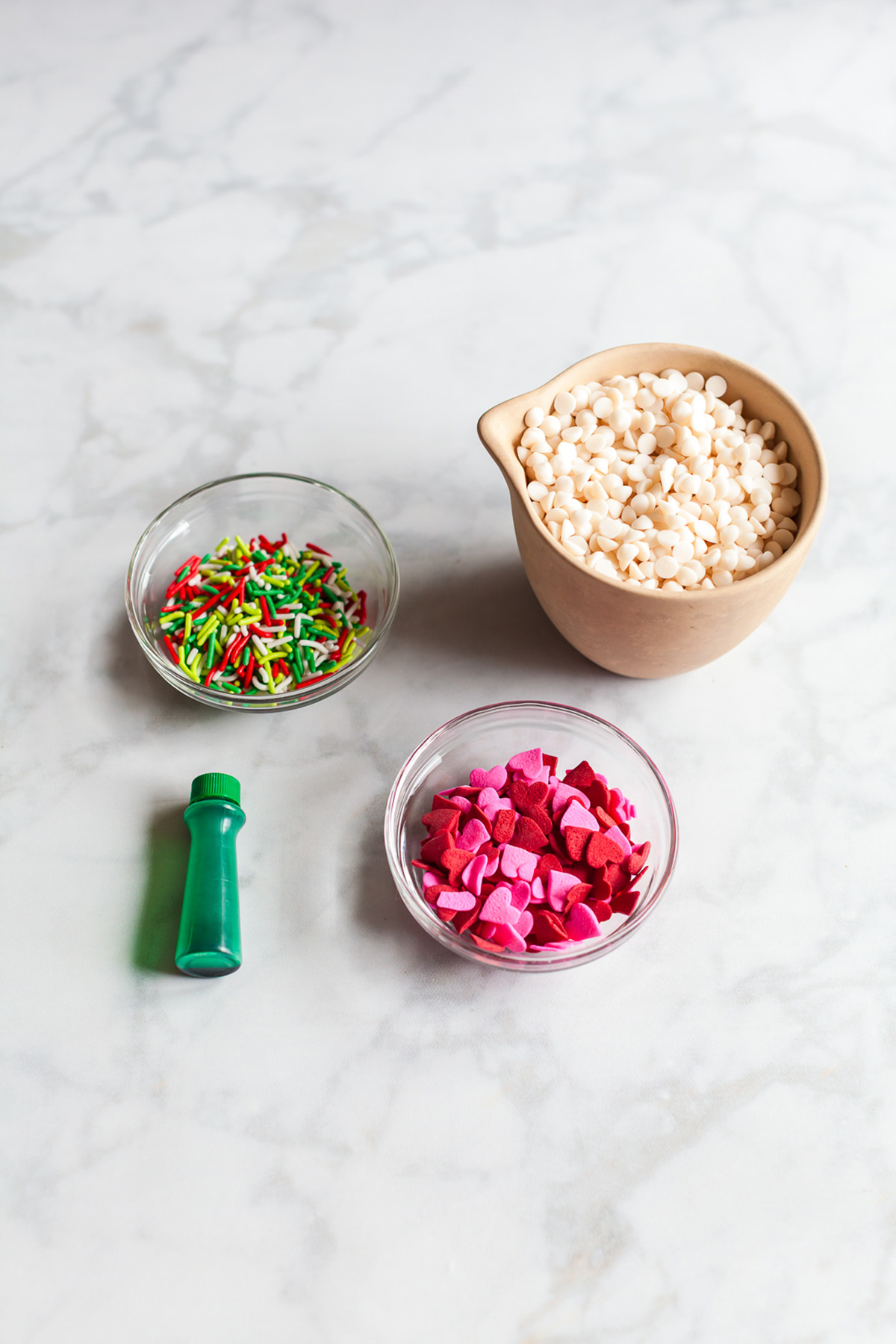 Bowls of white chocolate chips, heart sprinkles, and colorful jimmies arranged on a marble surface next to a bottle of green food coloring.