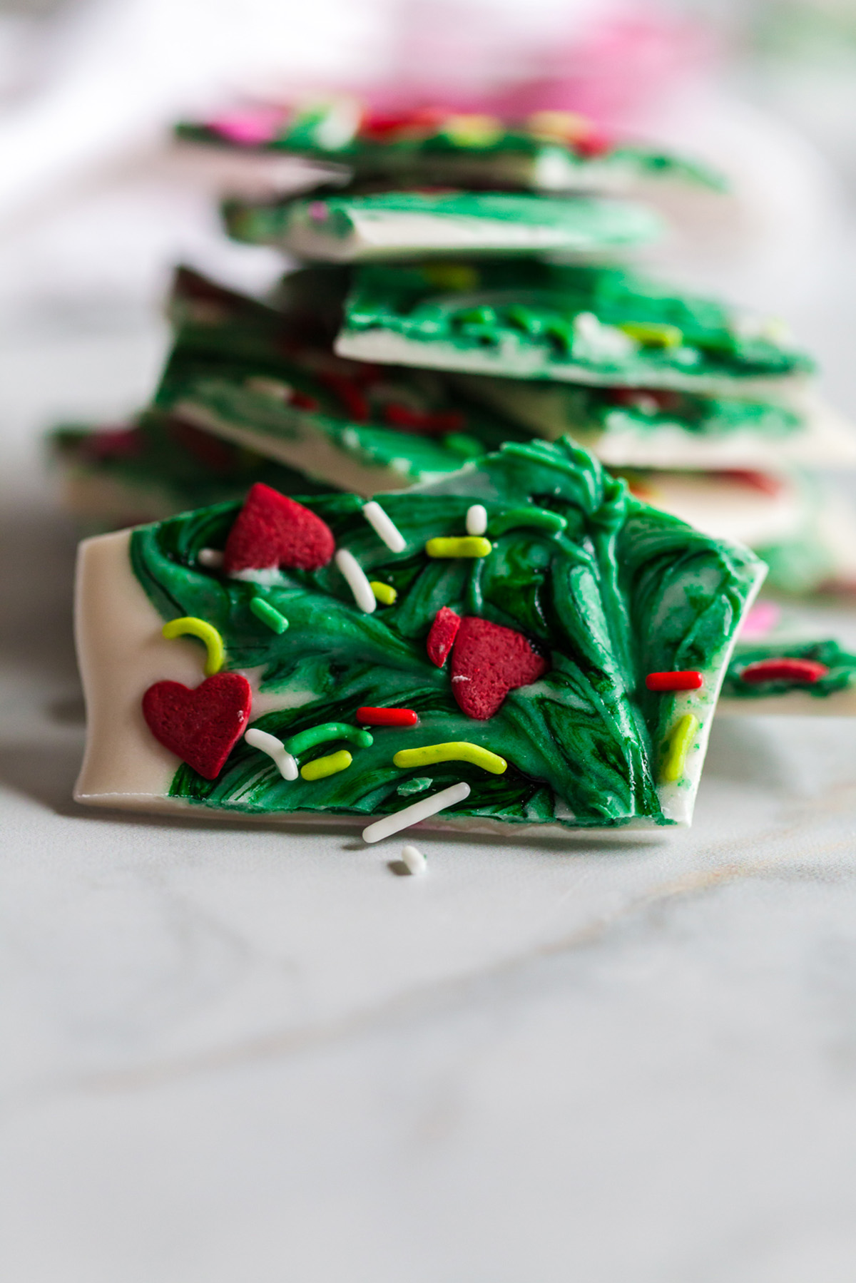 A close up of a piece of Grinch bark showing detailed green chocolate swirls, red candy hearts, and colorful sprinkles with a blurred stack of bark pieces behind it.