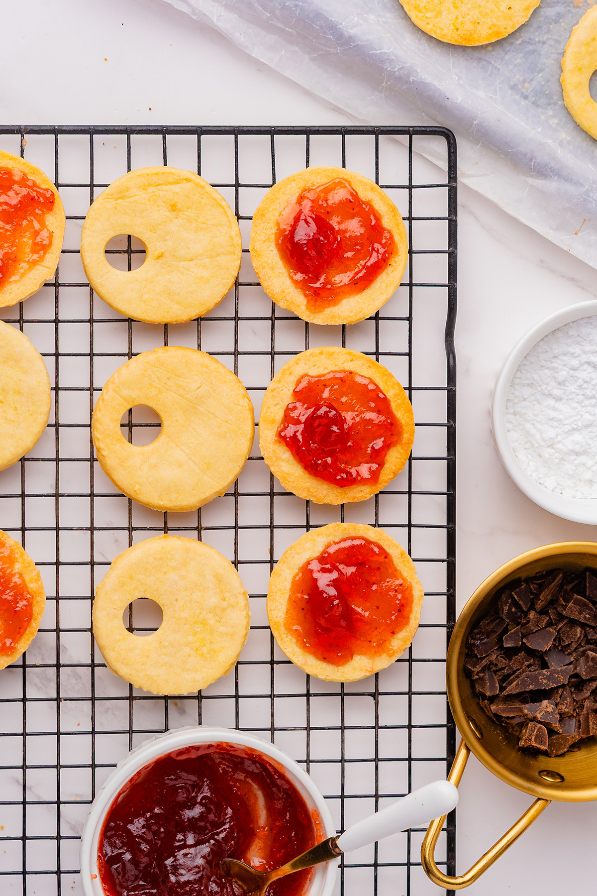 Linzer cookie rounds on a cooling rack with strawberry jam spread on the base cookies for assembling reindeer linzer cookies.