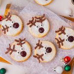Overhead view of several reindeer linzer cookies arranged on parchment paper with festive holiday ornaments.