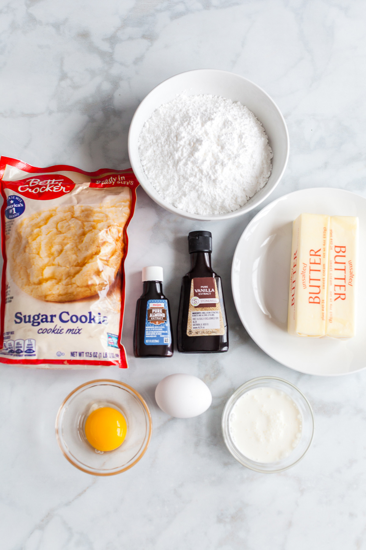Betty Crocker sugar cookie mix, butter, eggs, powdered sugar, vanilla extract, almond extract, and heavy cream on a marble countertop.