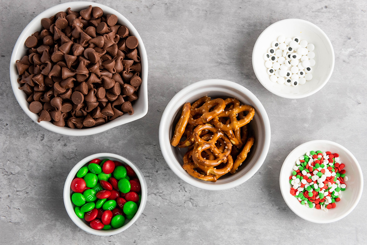 Bowls of ingredients for chocolate reindeer bark including milk chocolate chips, mini pretzels, red and green M&M’s, candy eyes, and festive sprinkles.