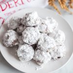 Close-up of powdered sugar–coated double chocolate snowball cookies piled on a white plate.