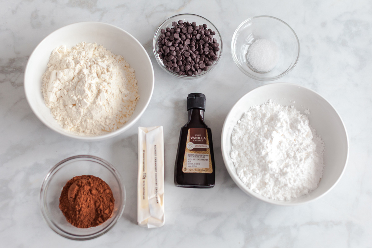 Flatlay of the ingredients for double chocolate snowball cookies, including flour, cocoa powder, butter, powdered sugar, vanilla, salt, and mini chocolate chips.