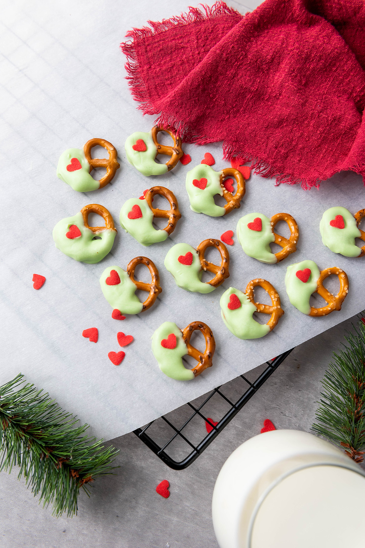 Finished Grinch pretzels cooling on parchment paper over a wire rack, surrounded by red sprinkles, a red cloth, pine greenery, and a glass of milk.