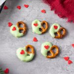 Green candy-coated pretzels with red heart sprinkles scattered on a gray backdrop with a red cloth and decorative hearts.