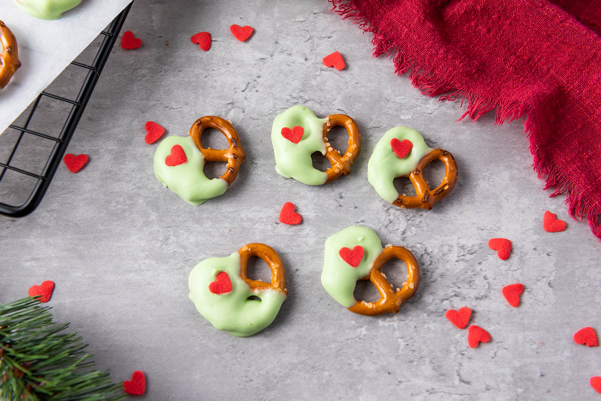 Green candy-coated pretzels with red heart sprinkles scattered on a gray backdrop with a red cloth and decorative hearts.