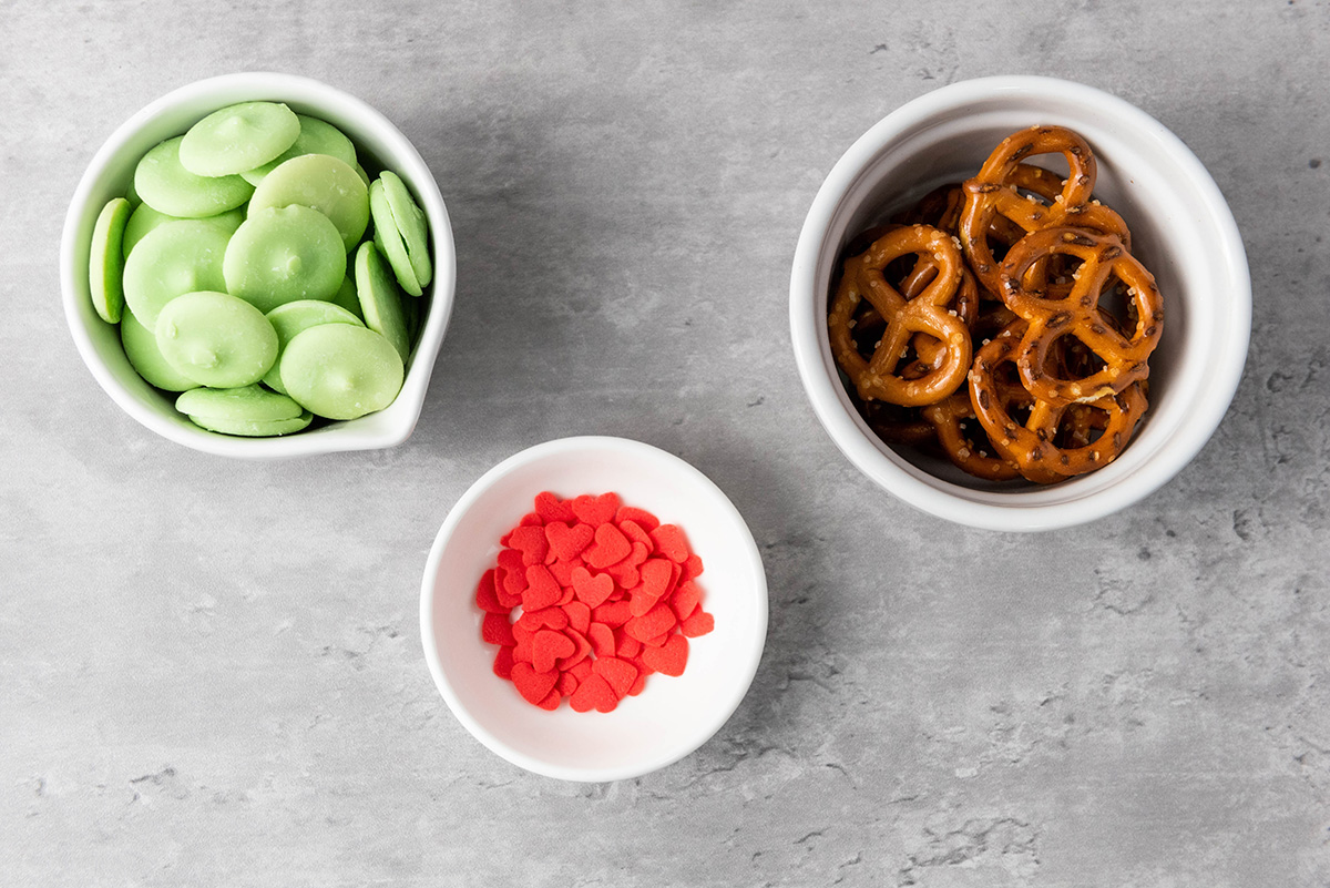 A top-down view of small bowls filled with green candy melts, mini pretzel twists, and red heart sprinkles arranged on a gray countertop.