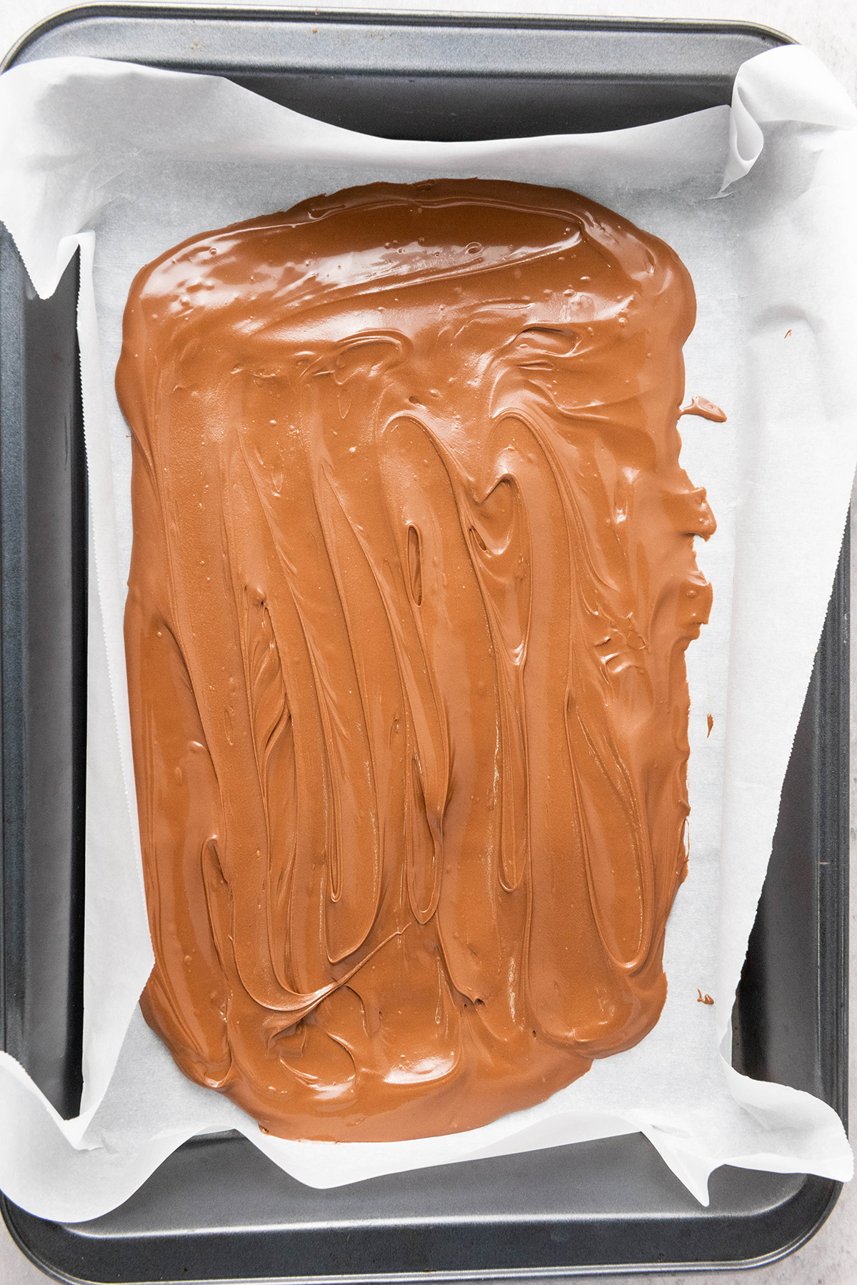 Smooth melted milk chocolate spread evenly in a parchment-lined baking pan, ready for toppings.