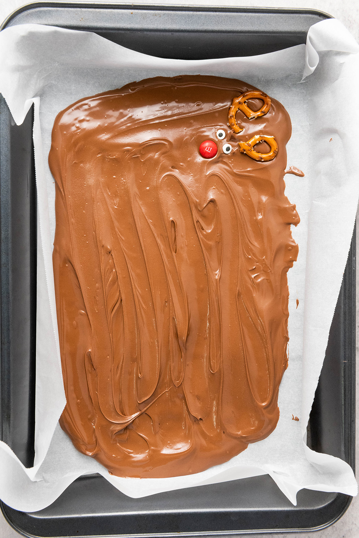 Melted milk chocolate spread in a lined baking pan with one reindeer face started in the top corner using pretzels, candy eyes, and a red M&M.