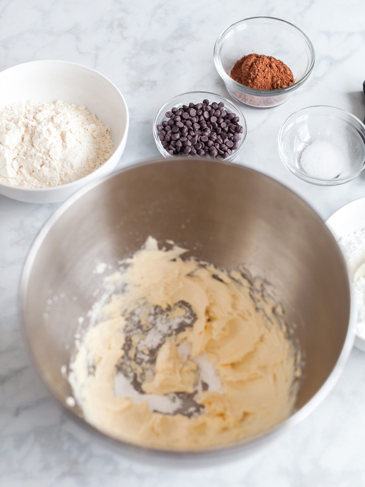 Creamed butter and powdered sugar in a mixing bowl with bowls of flour, cocoa powder, chocolate chips, and salt arranged around it.