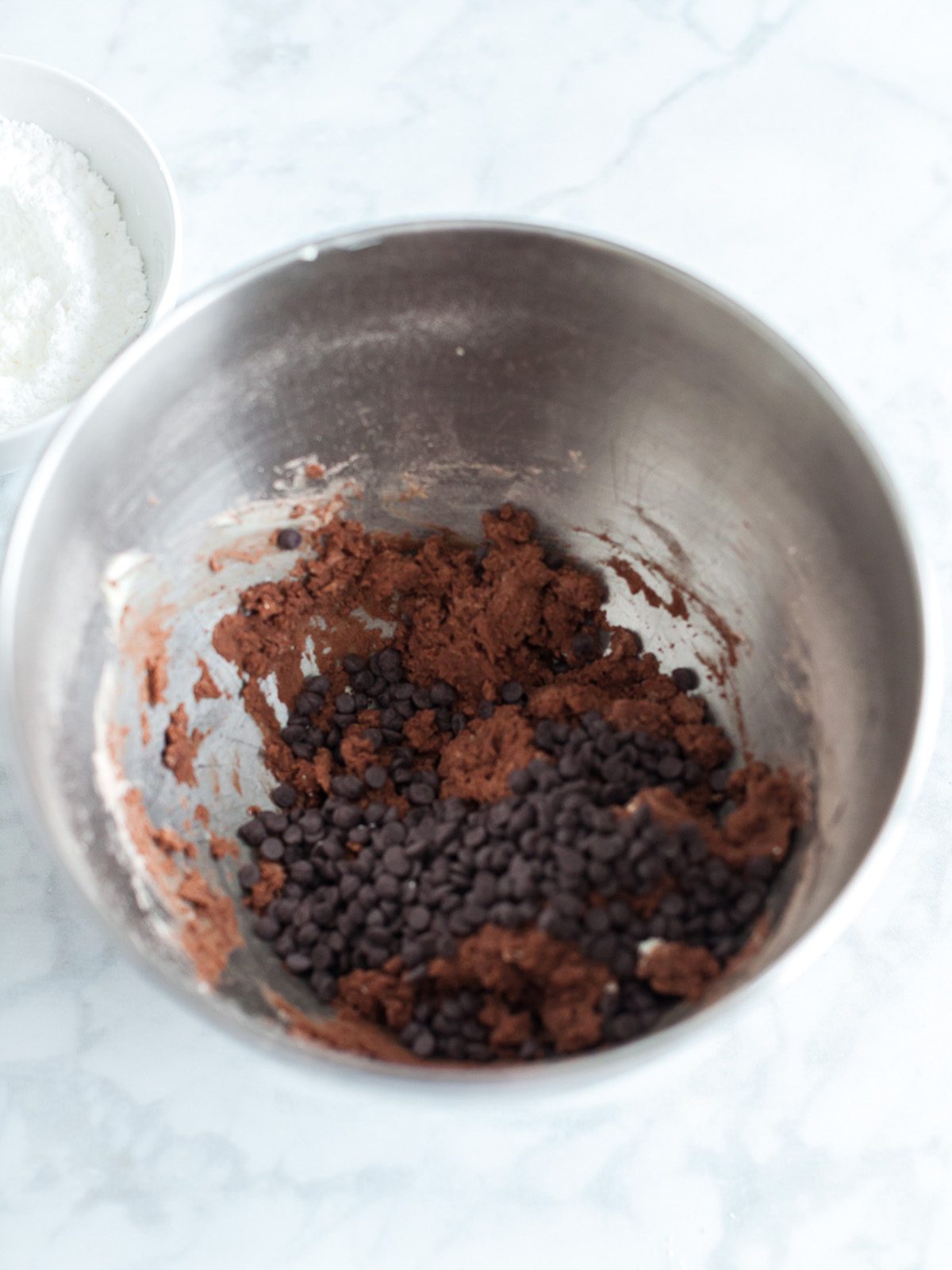 Chocolate cookie dough forming in a mixing bowl with cocoa powder mixed in and mini chocolate chips added on top.