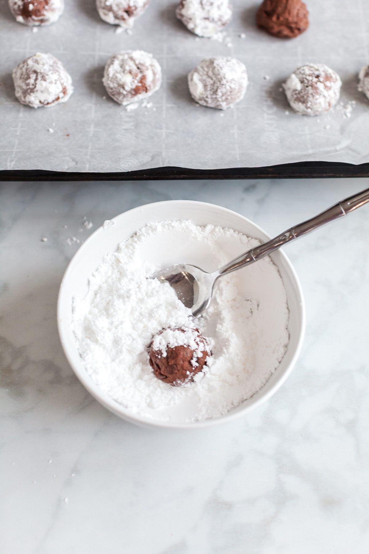 Warm chocolate cookie ball being rolled in a bowl of powdered sugar with a spoon.