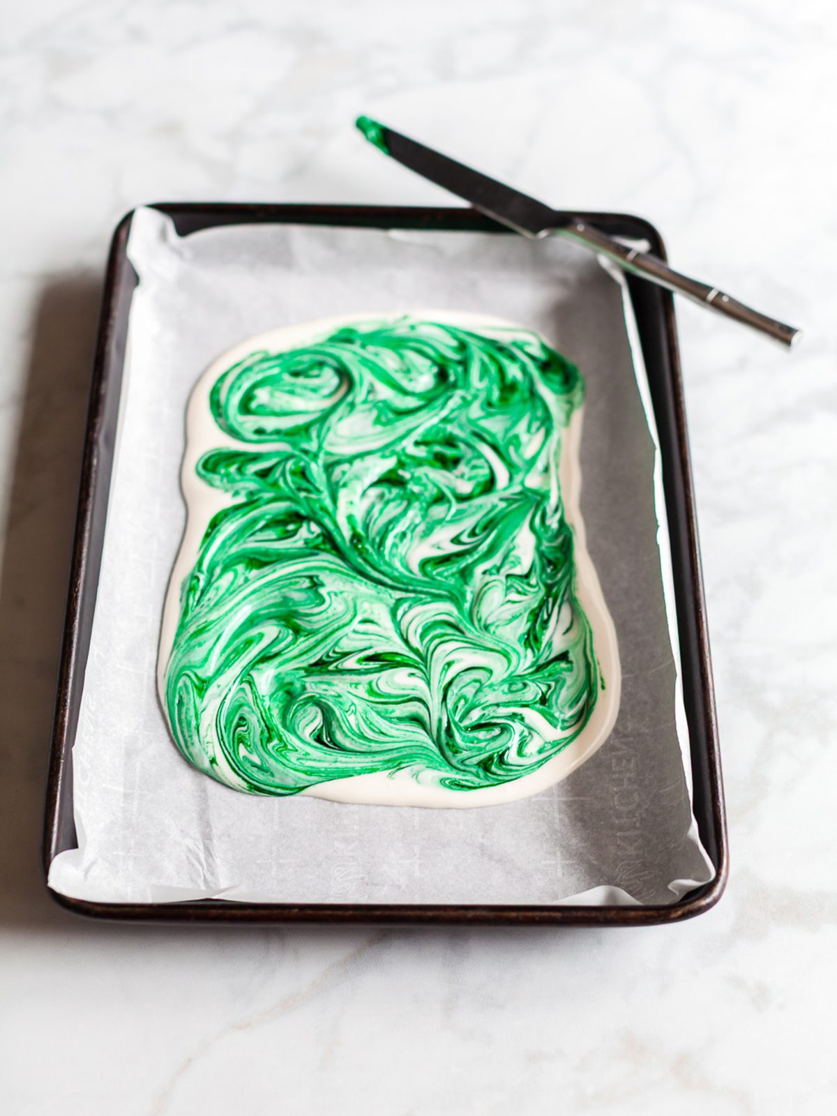 Freshly swirled white and green colored chocolate spread on a parchment lined sheet pan with a knife resting on the edge.