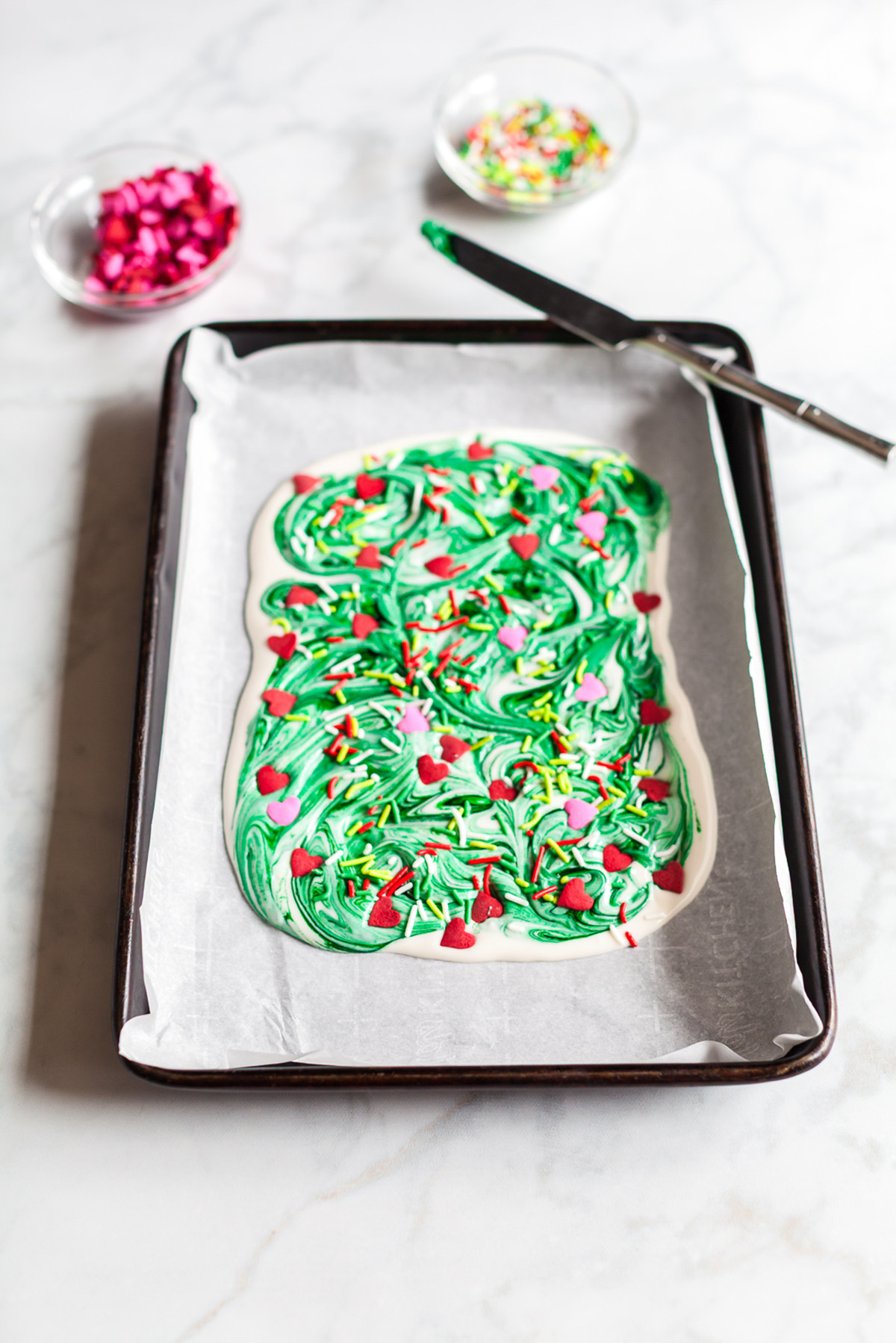 White chocolate bark swirled with bright green food coloring and topped with heart sprinkles and Christmas jimmies on a parchment lined sheet pan, with small bowls of sprinkles in the background.
