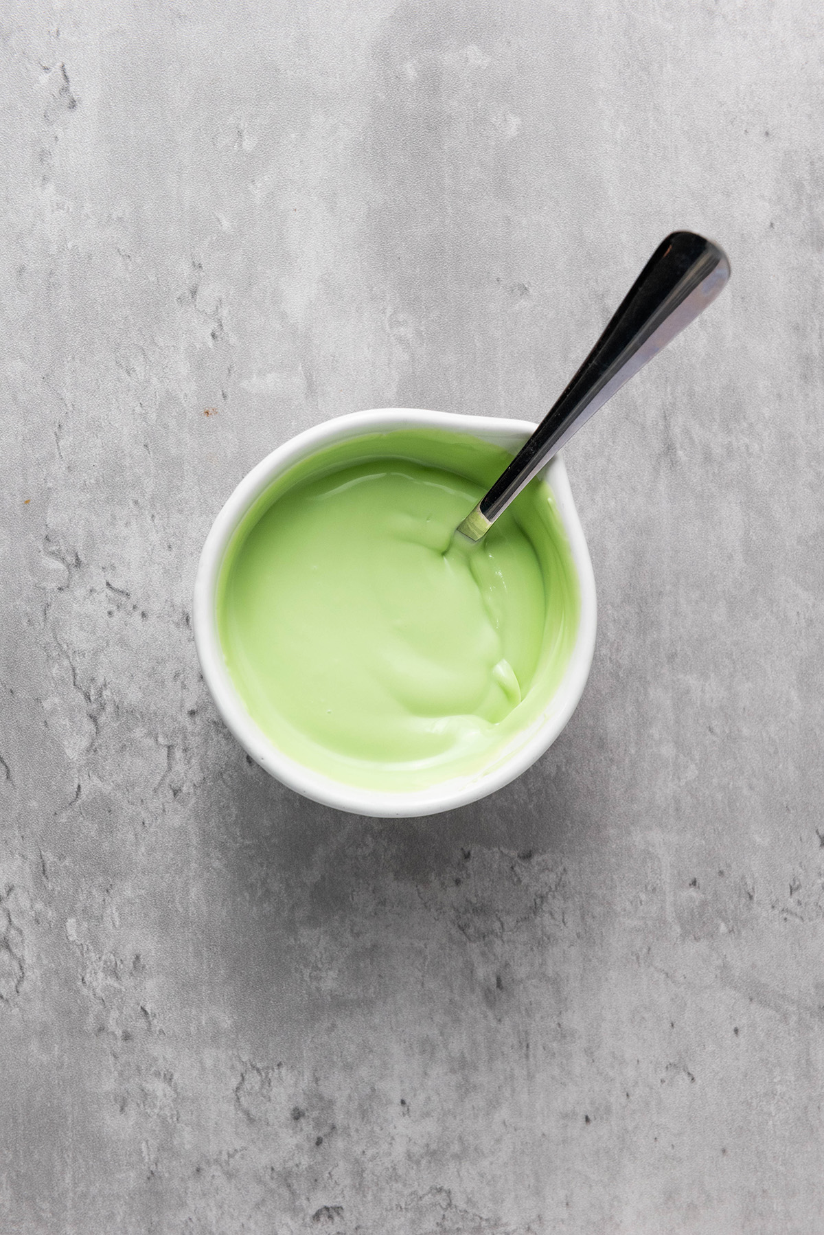 A small white bowl filled with smooth melted green candy melts, stirred with a spoon, sitting on a gray surface.