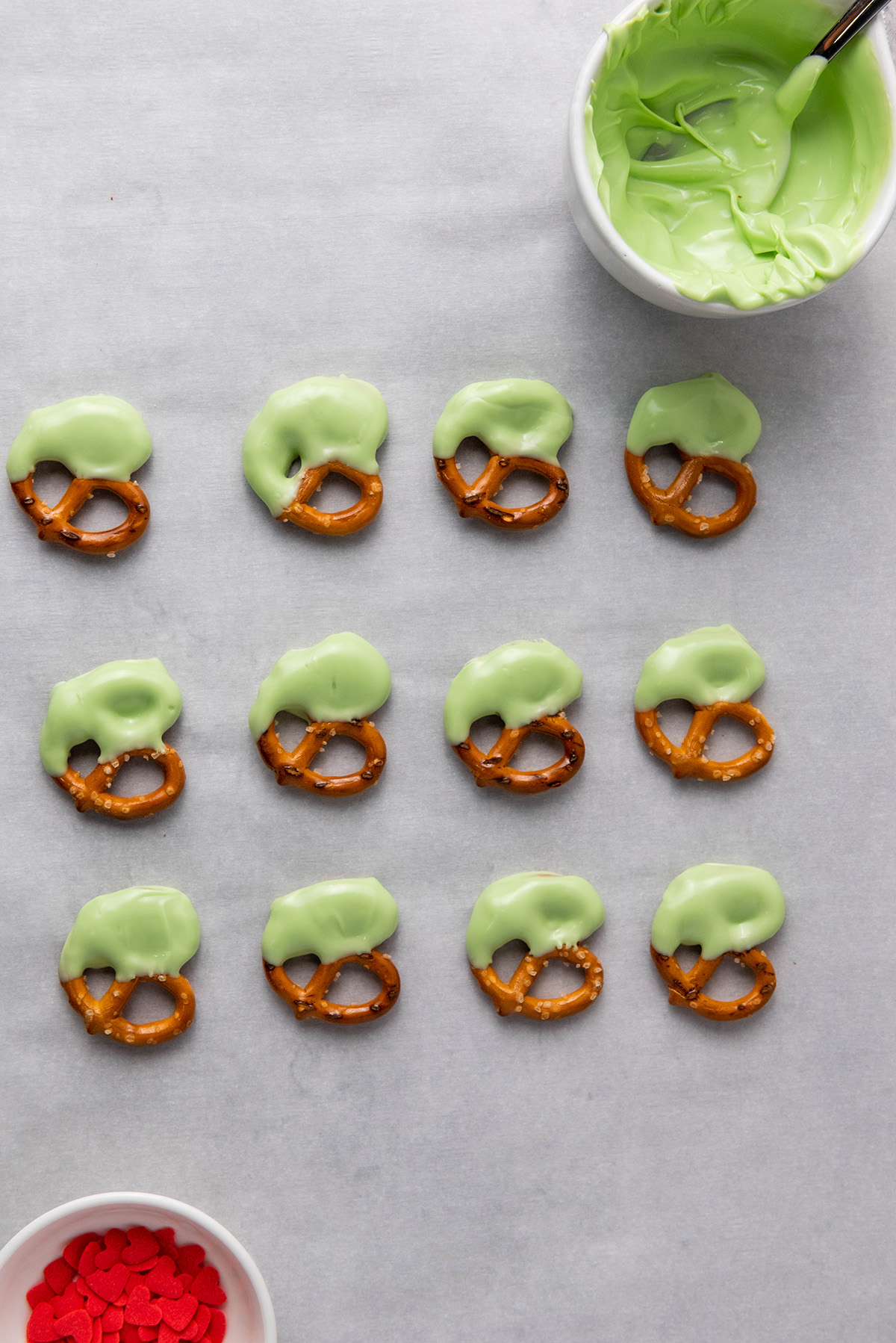 Mini pretzel twists partially dipped in melted green candy and lined up on a parchment-covered baking sheet.
