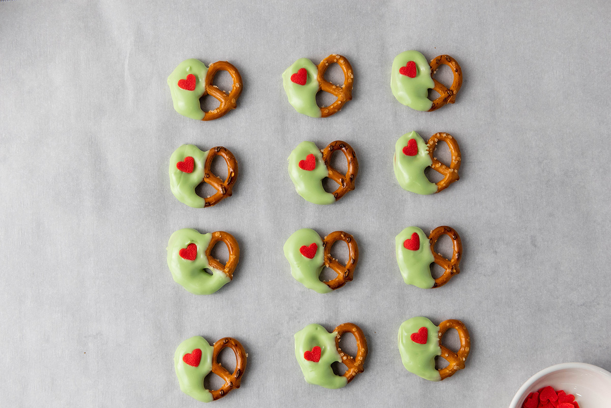 Partially dipped green pretzels topped with small red heart sprinkles, arranged neatly on parchment paper.