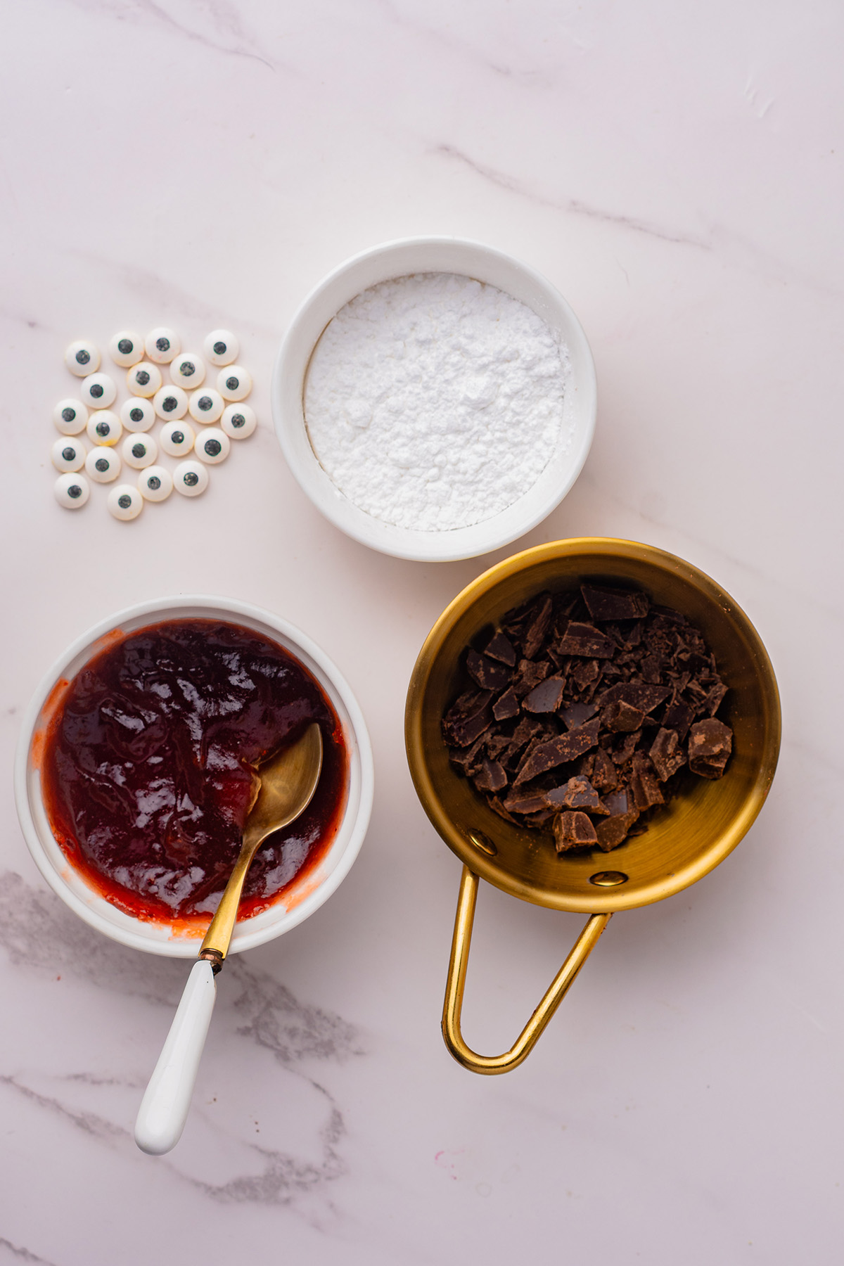 Candy eyeballs, powdered sugar, strawberry jam, and chopped chocolate displayed in bowls for decorating reindeer linzer cookies.