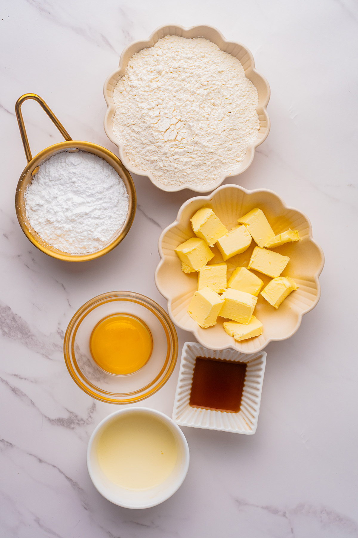 Bowls of flour, powdered sugar, cubed butter, egg yolk, heavy cream, and vanilla extract arranged on a marble surface for making reindeer linzer cookies.