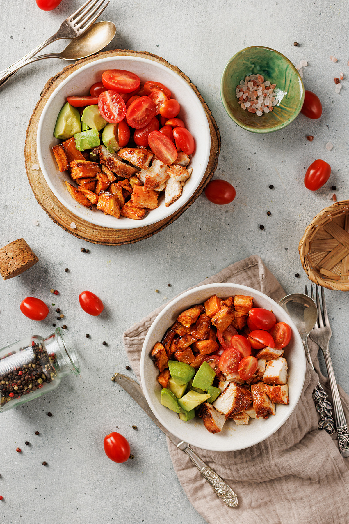 Blackened chicken and sweet potato bowls made with bold spices, roasted sweet potatoes, fresh tomatoes, and creamy avocado. Easy and satisfying. Two blackened chicken and sweet potato bowls styled on a table with fresh tomatoes, avocado, and utensils.