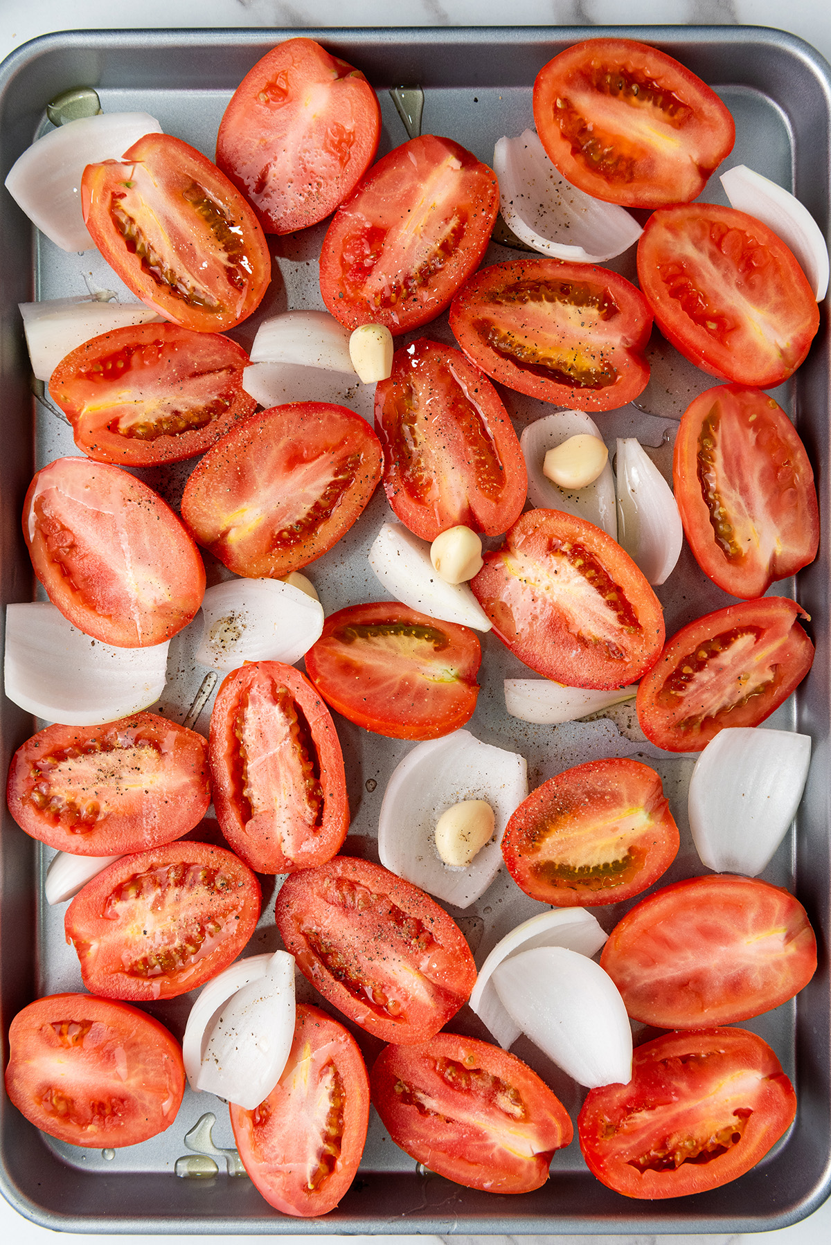 Halved Roma tomatoes, onion wedges, and garlic cloves arranged on a baking sheet before roasting.