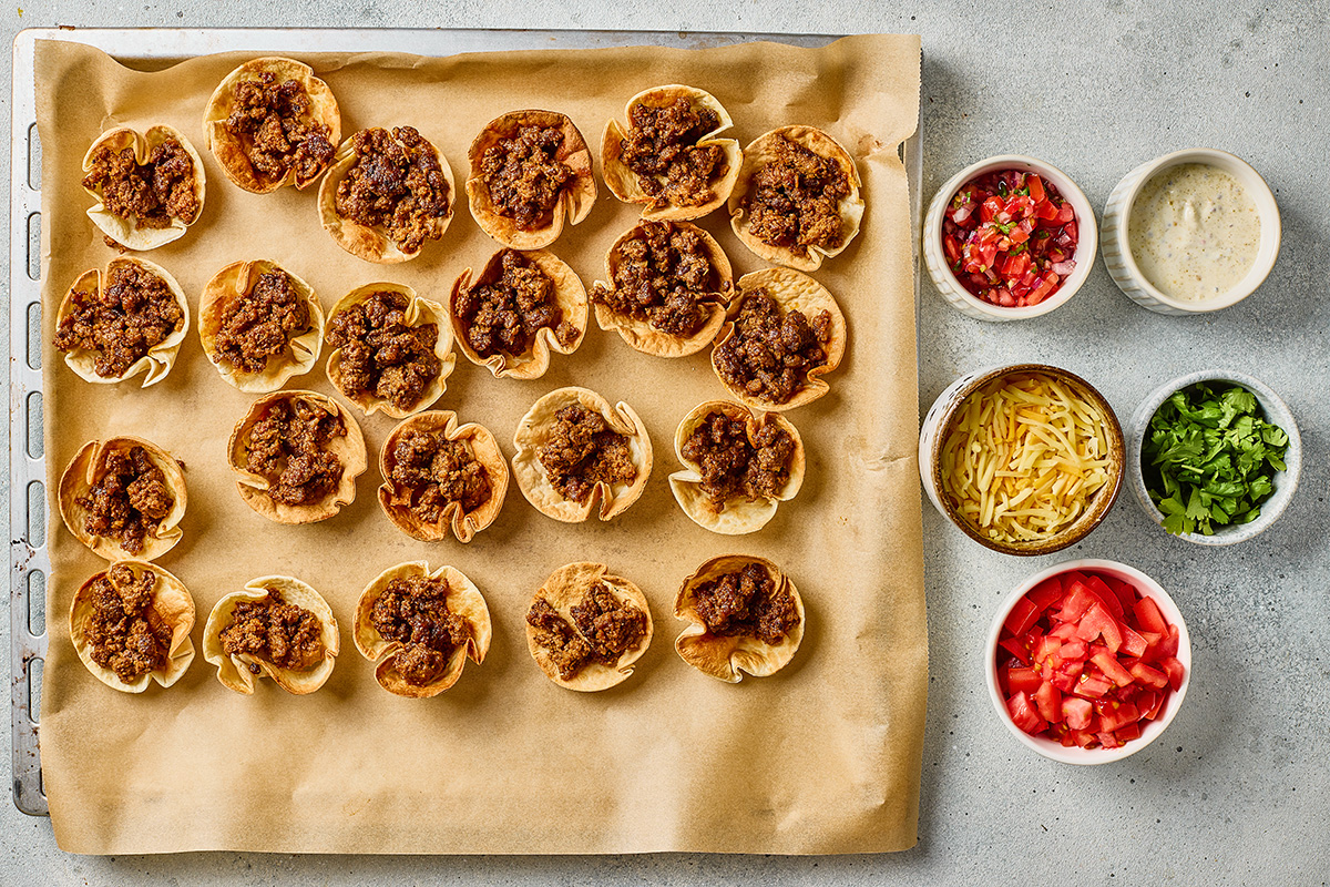 Tortilla scoop chips filled with seasoned taco beef arranged on a parchment-lined baking sheet.