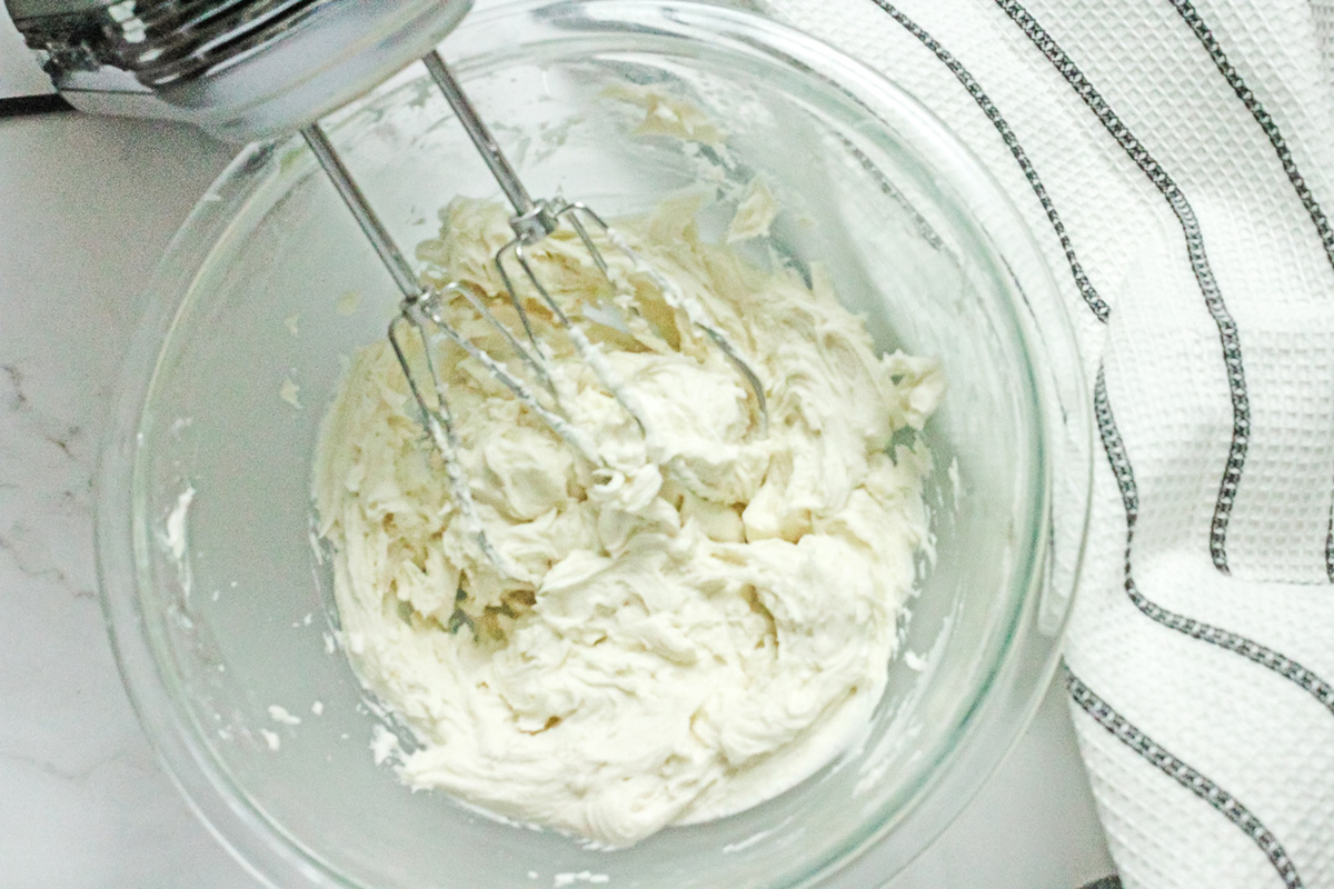 Vanilla frosting being whipped with a hand mixer in a glass bowl.