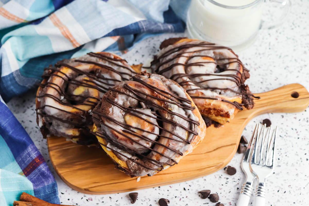 Chocolate brownie batter stuffed cinnamon rolls with icing and chocolate drizzle served on a wooden board with a glass of milk.