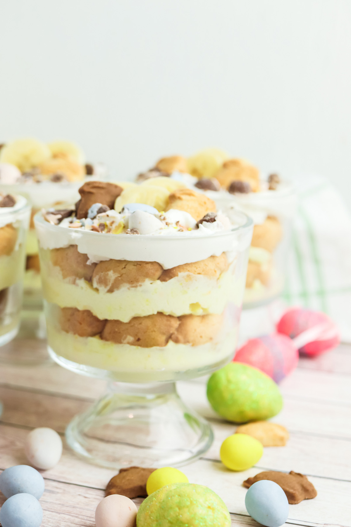 Single banana pudding cup in a glass trifle jar with visible layers, topped with bunny cookies and banana slices, with pastel eggs nearby.