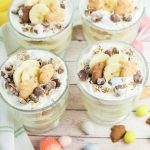 Four Easter banana pudding cups on a wood table with a green striped towel, bananas in the background, and pastel candy eggs scattered around.
