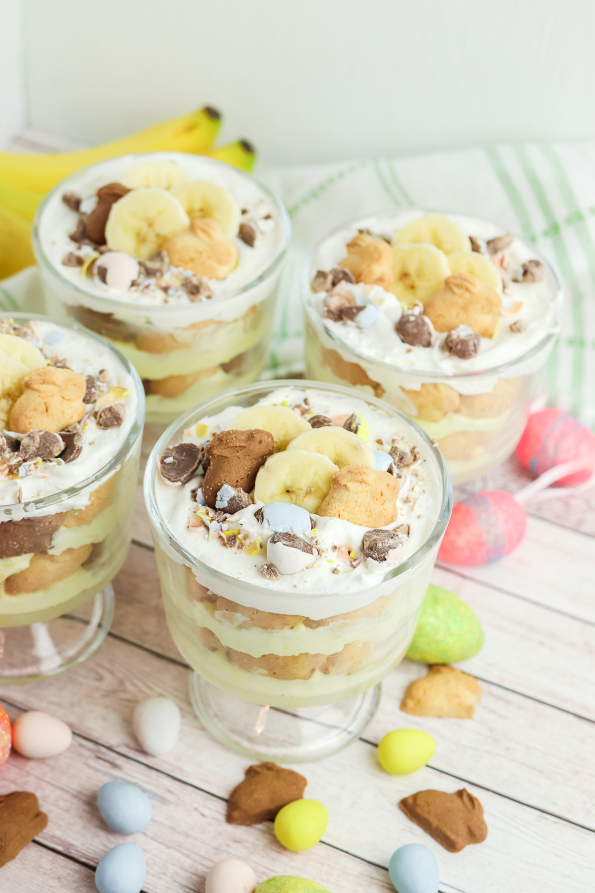 Overhead view of four layered banana pudding cups topped with banana slices, bunny cookies, crushed mini eggs, and chocolate chunks.