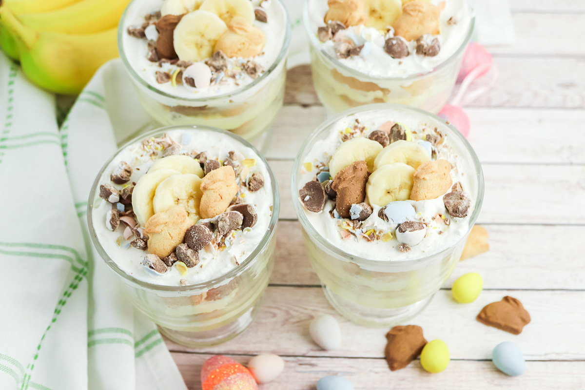 Four Easter banana pudding cups on a wood table with a green striped towel, bananas in the background, and pastel candy eggs scattered around.