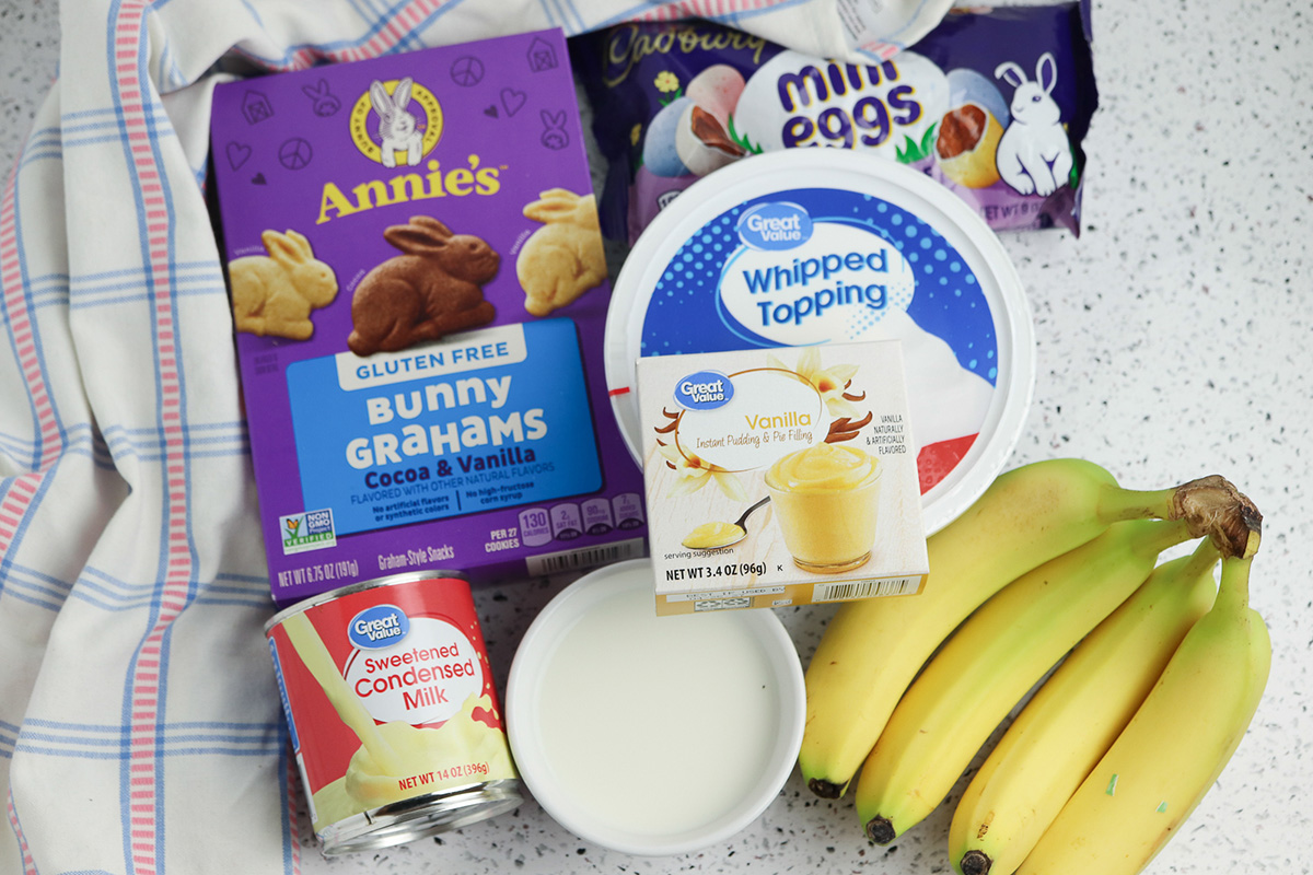 Ingredients for Easter banana pudding cups including bananas, instant vanilla pudding mix, sweetened condensed milk, whipped topping, bunny cookies, and mini eggs.