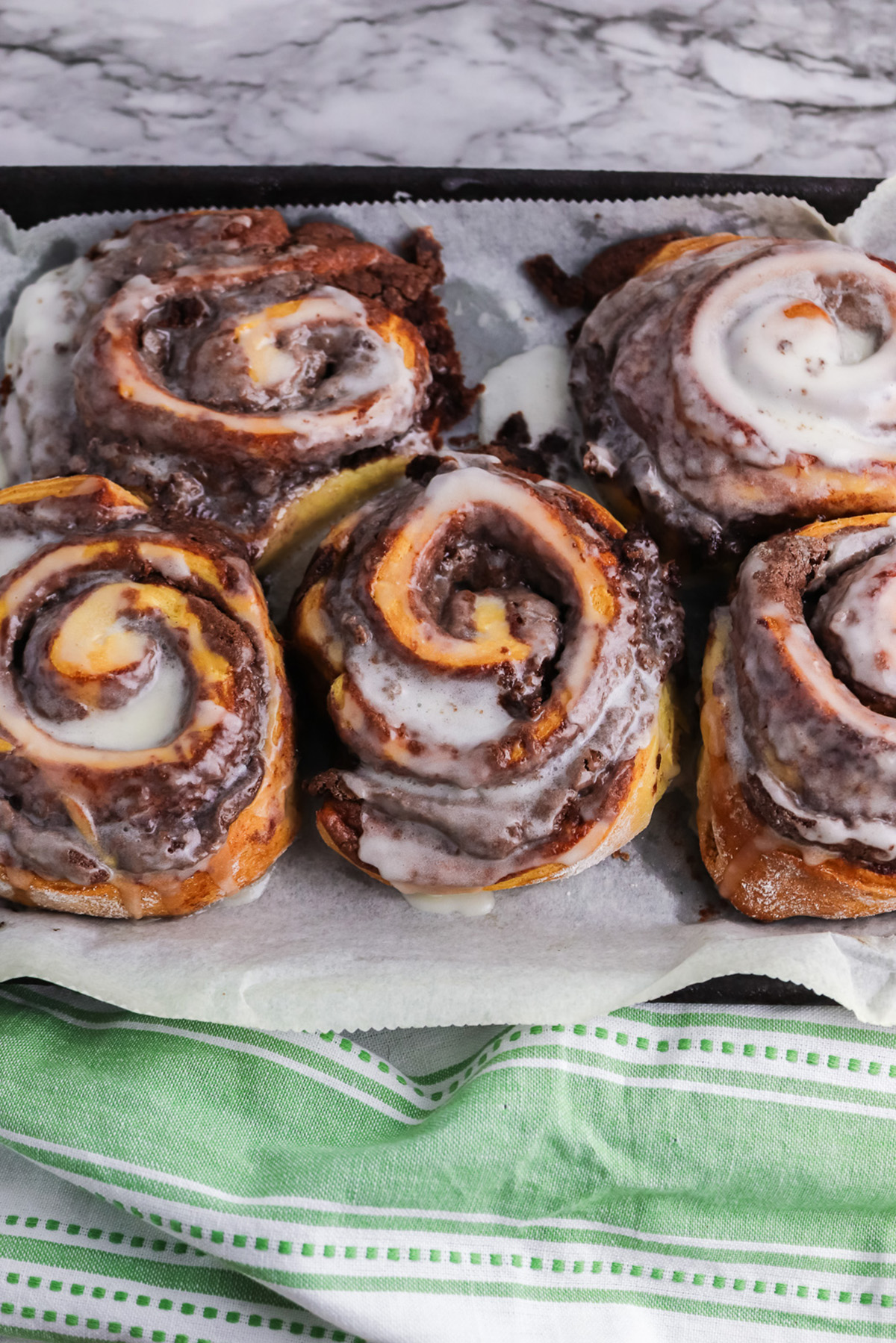 Brownie cinnamon rolls baked on parchment paper with icing melting over the warm swirls.