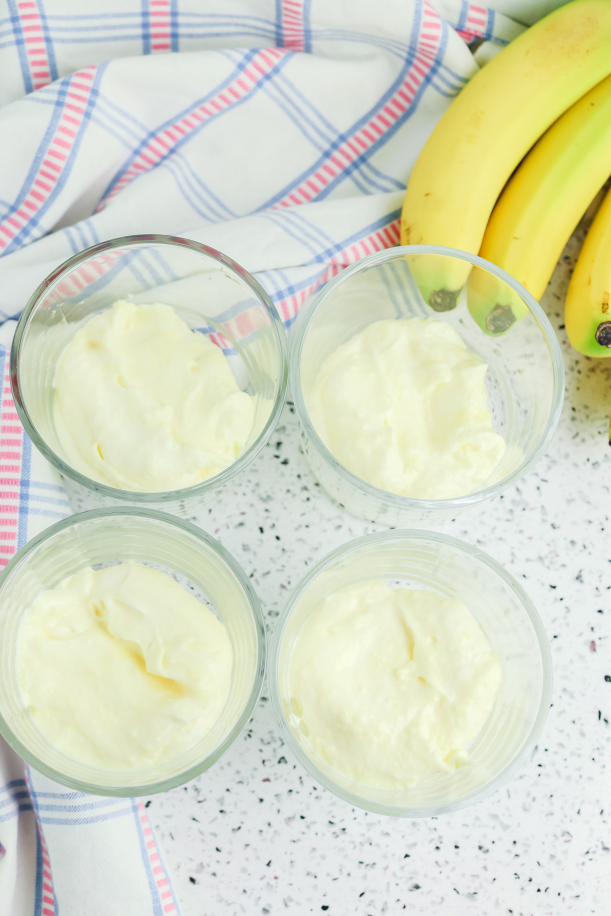 Four dessert cups filled with vanilla pudding mixture, with bananas and a plaid kitchen towel in the background.