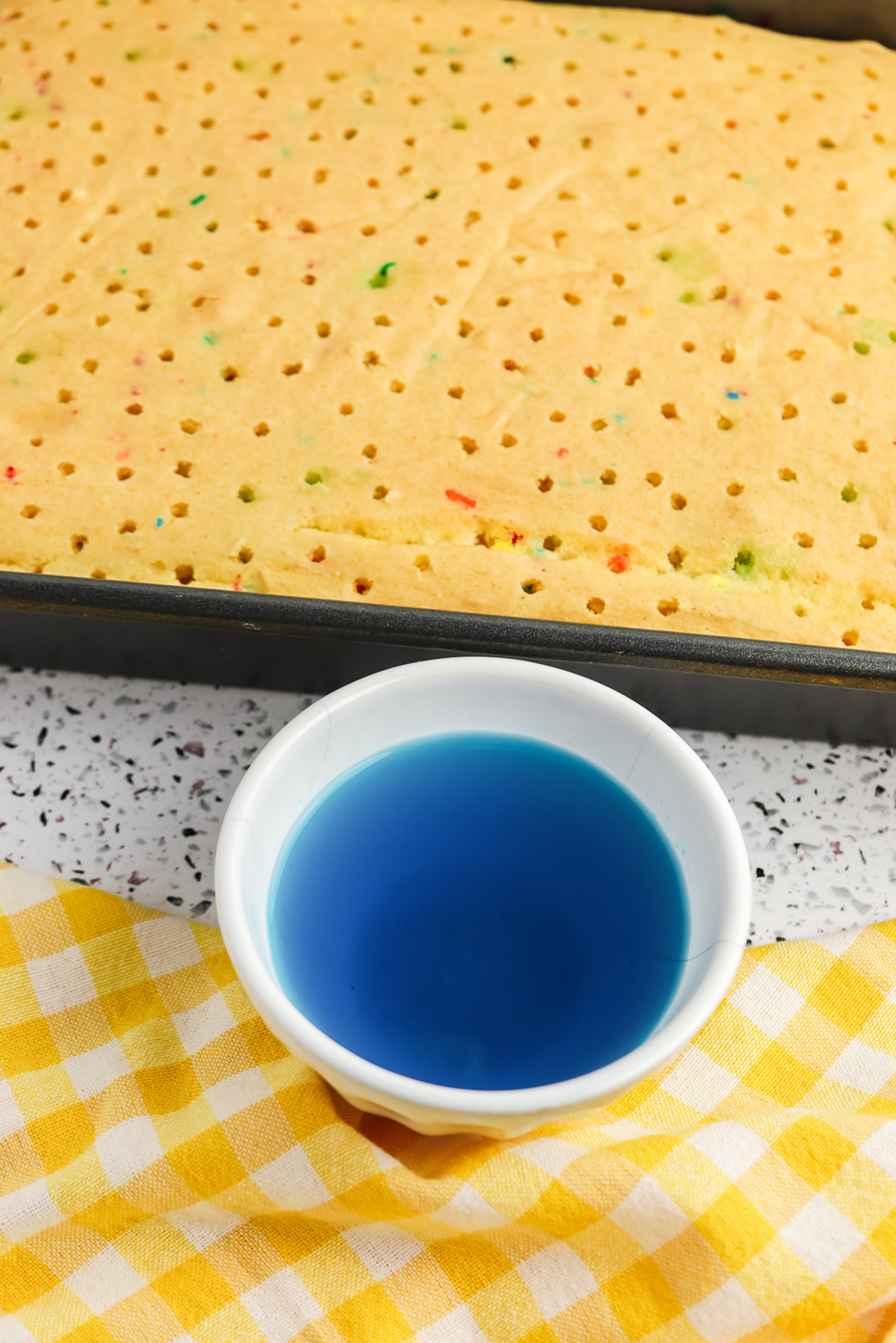 Poked Funfetti cake in a pan with a bowl of blue Jello mixture for rainbow poke cake.