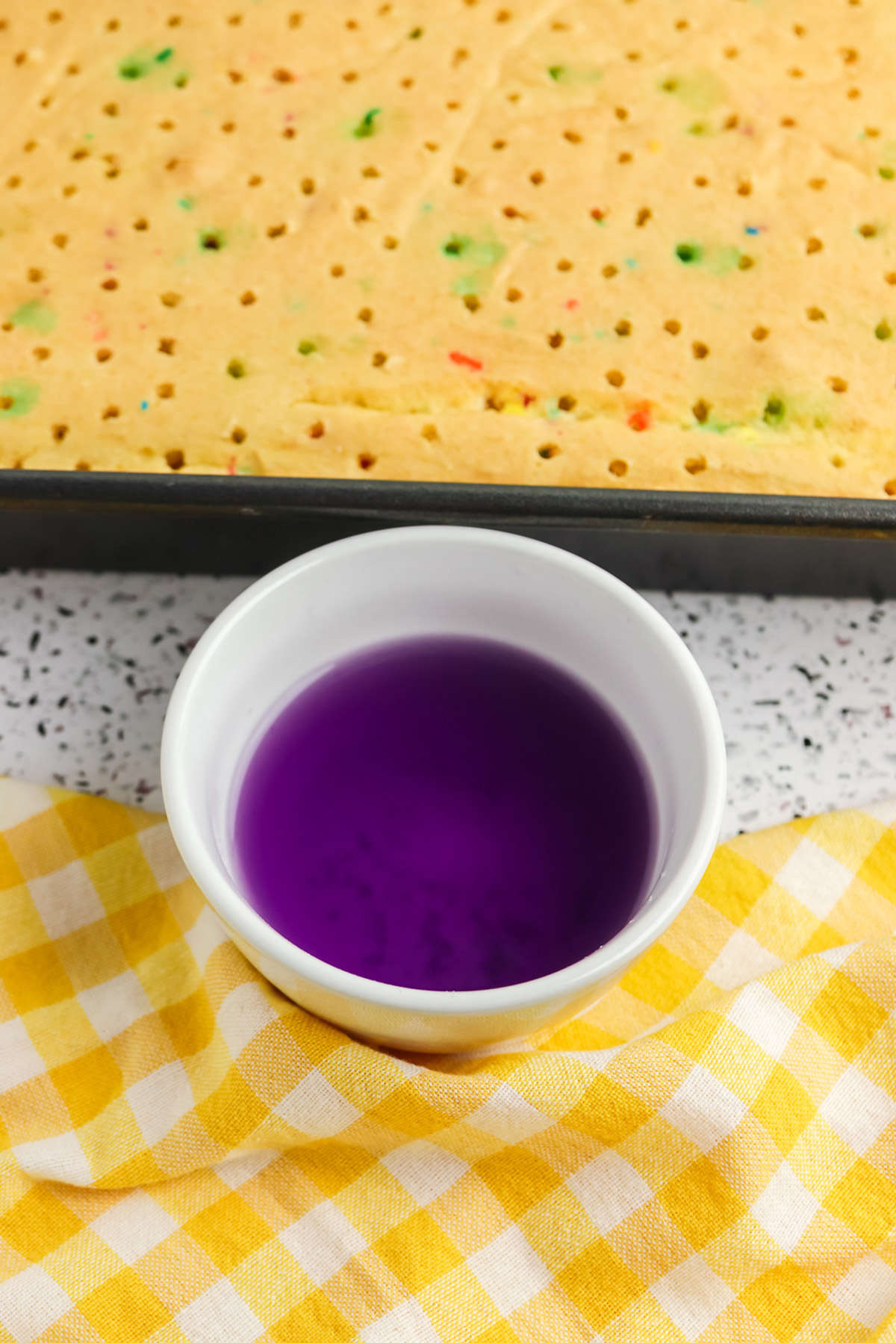 Poked Funfetti sheet cake with a bowl of purple Jello mixture for making rainbow Jello poke cake.