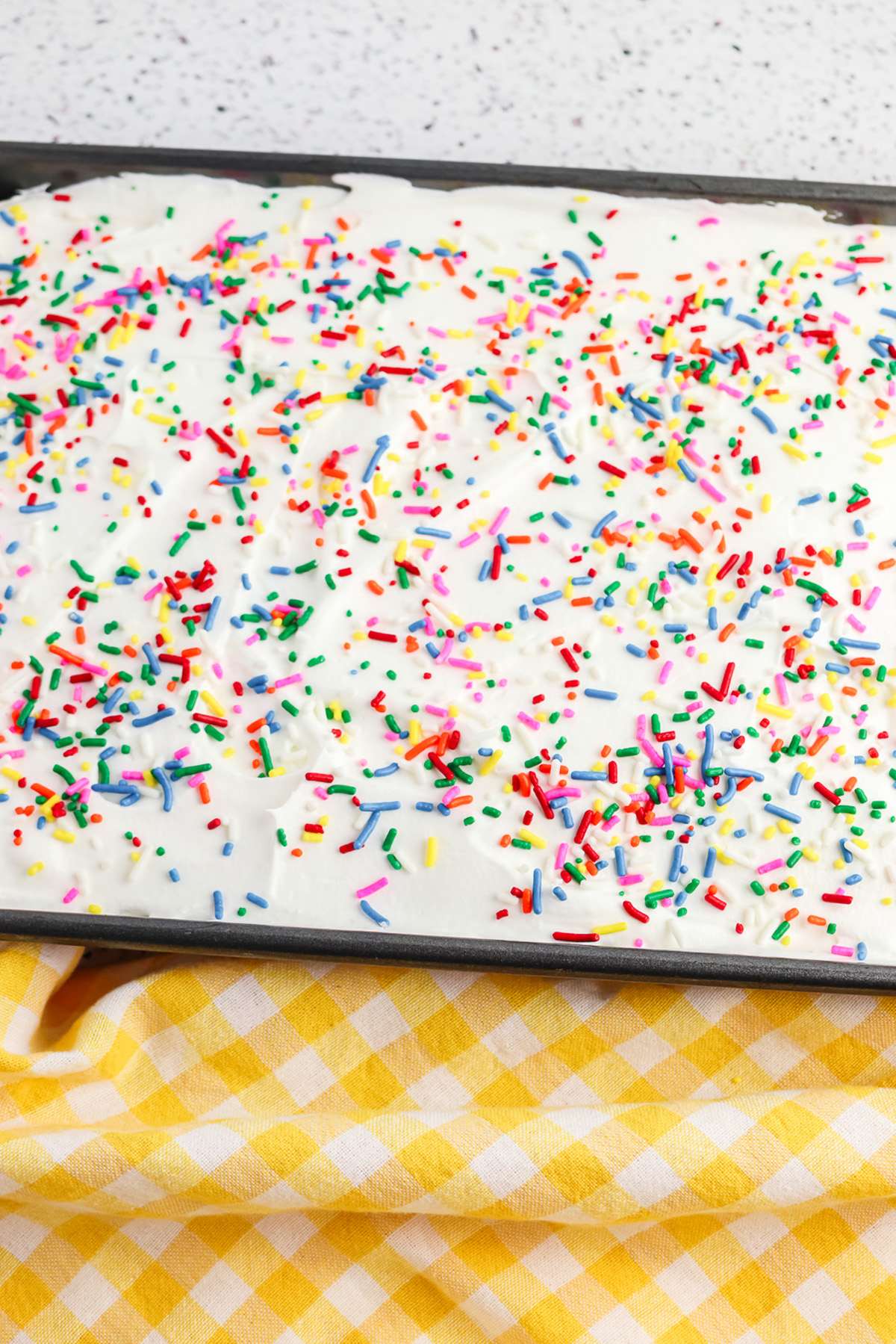 Rainbow poke cake frosted with whipped topping and covered with rainbow sprinkles in a baking pan.