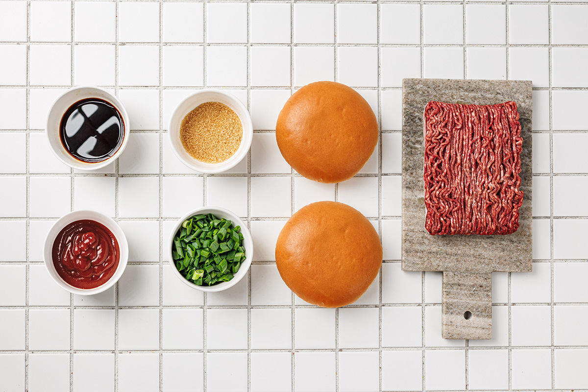 Korean sloppy joes ingredients including ground beef, soy sauce, brown sugar, ketchup, sesame oil, brioche buns, green onions, and sesame seeds arranged on a white tile background.